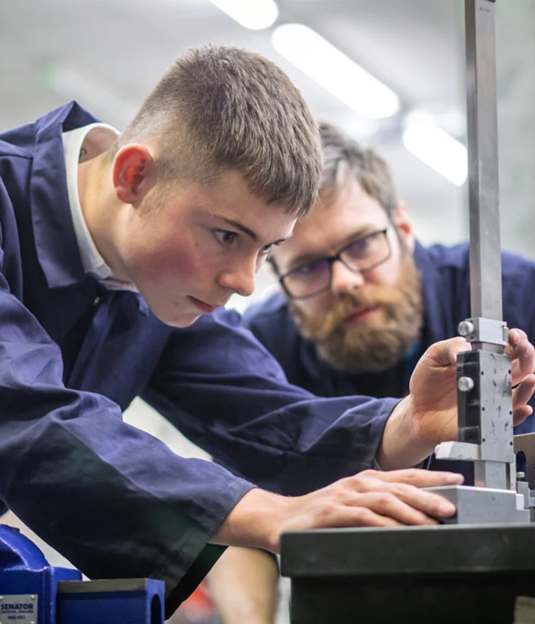 A lecturer and a student in navy-blue overalls working together in a mechanical engineering workshop. The lecturer is demonstrating how to use a tool on a metal component held in a vise. A lecturer and a student in navy-blue overalls working together in a mechanical engineering workshop. The lecturer is demonstrating how to use a tool on a metal component held in a vise.